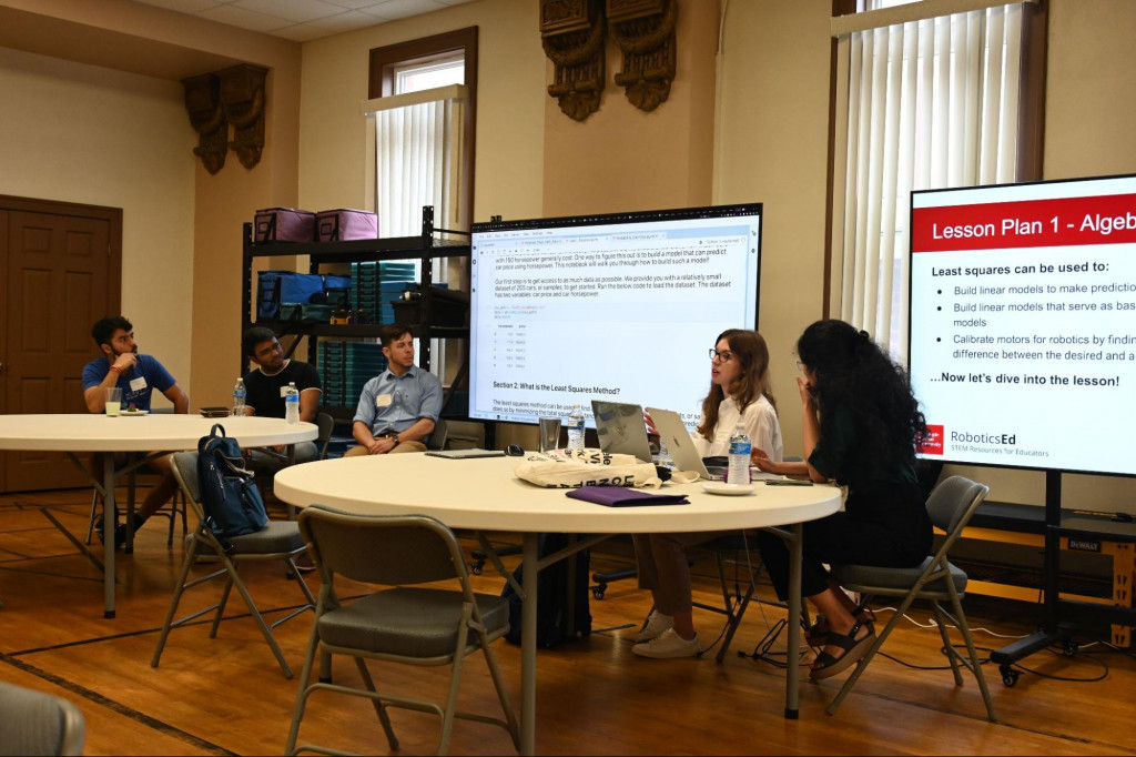Five members of the RoboticsEd team are seated at two large circular tables in front of two large screens to present their interactive lesson plans on core mathematical concepts and how they relate to Robotics Research.