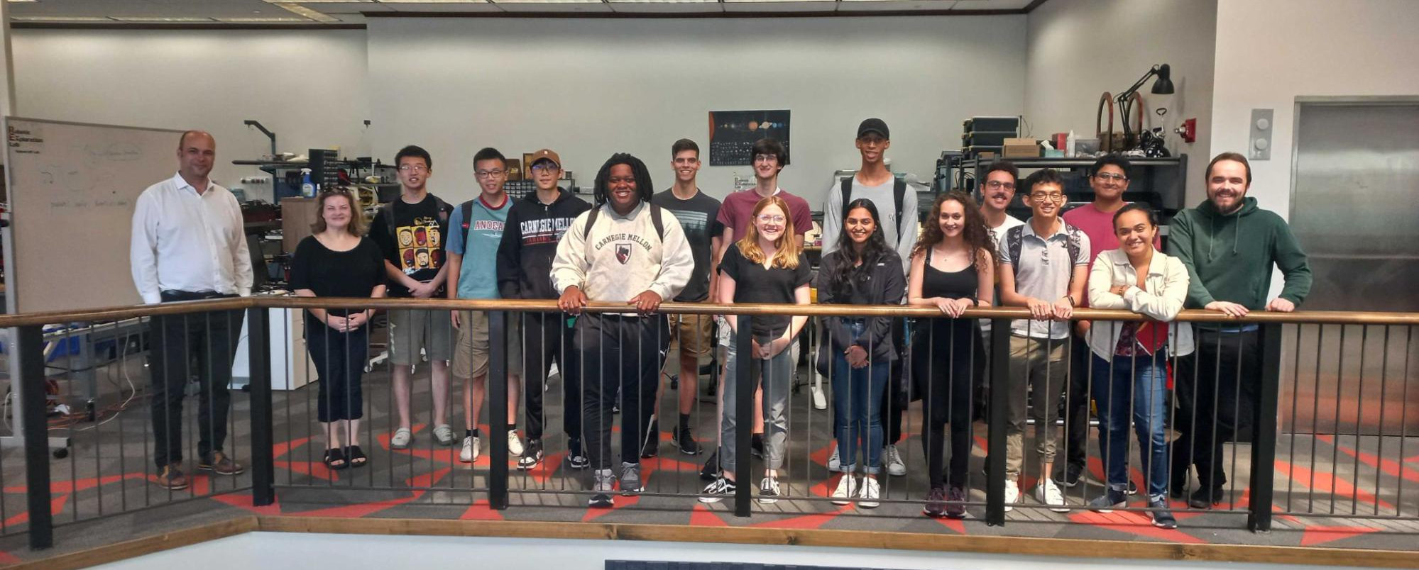 Fifteen students posed in a lab with the front row leaning over a wooden banister. The head researcher and the program director are positioned the furthest left in the grouping. Lab equipment is partially visible behind the group.